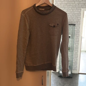 Grå stickad tröja från C.P. Company - Säljer en grå stickad tröja från C.P. Company med rund halsringning och en bröstficka med knappdetaljer. Tröjan har långa ärmar och ribbade muddar vid ärmslut och nederkant. Perfekt för lager-på-lager-stil.