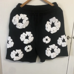 Svarta shorts med vita blommor från Denim Tears - Svarta shorts från Denim Tears med stora vita blommor som mönster över hela plagget. Shortsen har resår i midjan med snörning och en bakficka. Perfekta för en avslappnad och trendig stil. Men utan organal förpackning 