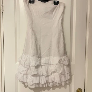 Vit volangklänning från VILA - Säljer en söt vit klänning från VILA med flera volanger nertill. Axelbandslös modell med kort längd, perfekt för sommarens tillfällen. Klänningen har en lätt glansig yta.