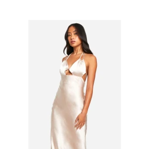 Champagnefärgad långklänning i satin med öppen rygg - Säljer en elegant champagnefärgad långklänning i satin med smala axelband och öppen rygg. Klänningen har en v-ringad omlott-topp och en figurnära passform som faller snyggt längs kroppen. Perfekt för speciella tillfällen! Balklänning