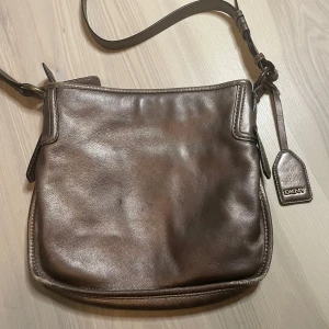 Bronsfärgad axelväska från DKNY - Snygg axelväska från DKNY i bronsfärgat skinn med justerbar axelrem och dragkedja upptill. Väskan har ett rymligt huvudfack med innerfickor och klassisk DKNY-tag på sidan. Perfekt för dig som vill ha en stilren och praktisk accessoar.
