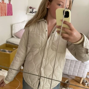 Beige quiltad jacka från SAMSA Young - Quiltad beige jacka från SAMSA Young med snygg vågig sömnad, tryckknappar och två bröstfickor. Jackan har klassisk krage och är långärmad, perfekt för lager-på-lager. Materialet är mjukt och lätt, passar till chill vardagsstil.