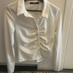 Vit långärmad skjorta från BikBok - Snygg vit skjorta från BikBok i storlek M. Skjortan har lång ärm, klassisk krage och knappar framtill. Snygg rynkdetalj längs knappraden ger en modern och trendig look. Perfekt att styla med jeans eller kjol.