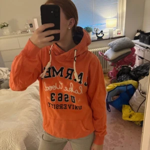 Orange hoodie  - Säljer en orange hoodie med broderad text framtill. Tröjan har huva med vita snören och en stor magficka. Perfekt för dig som gillar sportig och avslappnad stil.