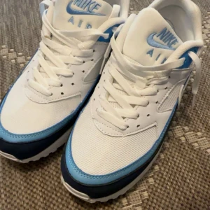 Nike Air Max sneakers vit/blå - Fräscha Nike Air Max sneakers i vitt och ljusblått med mörkblå detaljer. Klassisk mesh och syntet på ovandelen, vit sula med synlig Air-enhet och snörning. Perfekta för dig som vill ha en clean och sportig look med retrovibbar.
