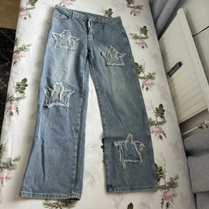 Blå jeans med stjärn-detaljer (aldrig använd) - Säljer ett par ljusblå jeans med raka ben och stora slitna stjärn-detaljer på benen. Jeansen har klassisk femficksdesign och är tillverkade i en mix av viskos och polyester. Passar för de med storlek S/M. 
