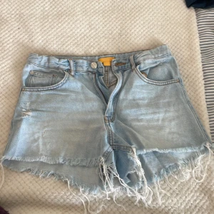 Ljusblå jeansshorts från Gina Tricot - Snygga ljusblå jeansshorts från Gina Tricot med hög midja och råa, fransiga kanter. Klassisk femficksmodell med slitningar framtill för en cool, avslappnad vibe. Perfekta för sommardagar och festivalhäng.
