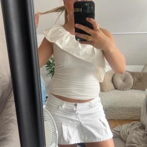 Vit oneshoulder topp med volang - Supersnygg vit topp med oneshoulder-design och stor volang över axeln. Tight passform som framhäver figuren och ger en trendig look. Perfekt för dig som vill sticka ut med en unik och stilren topp i sommar.