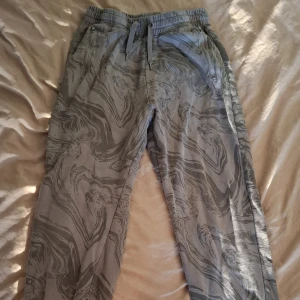 Grå mönstrade mjukisbyxor H&M - Säljer ett par grå mjukisbyxor från H&M i storlek 164. Byxorna har ett unikt marmorerat mönster, resår och snörning i midjan samt dragkedjefickor. Perfekta för chill dagar eller när du vill ha något bekvämt och snyggt.