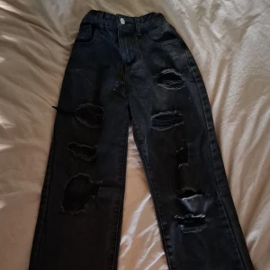 Svarta ripped jeans med raka ben - Säljer ett par svarta jeans med raka ben och flera stora slitningar framtill för en edgy look. Jeansen har klassisk femficksdesign och knappgylf. Perfekta för dig som gillar streetstyle och vill sticka ut.