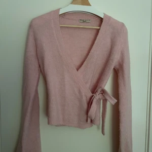 Rosa omlottkofta från GinaTricot - Supermjuk och fluffig rosa omlottkofta från Gina med knyt i sidan och v-ringad hals. Långärmad och i rosa färg, perfekt för lager-på-lager. Materialet är stickat och känns riktigt cozy. Använd fåtal gånger