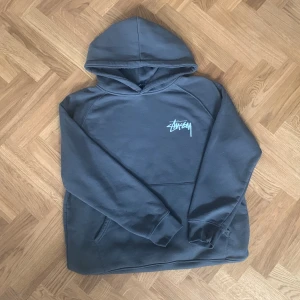 Grå Stüssy hoodie - Mörkgrå hoodie från Stüssy med stor vit logga på ryggen och mindre logga på bröstet. Klassisk känguruficka och huva. Mjuk bomullskänsla, perfekt för chill dagar. Långärmad och loose fit för en avslappnad streetstyle.