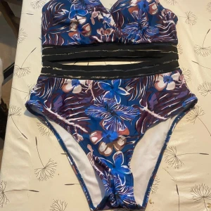 Blommig bikini med cut out-detaljer - Snygg bikini med blommigt mönster i blått, lila och beige. Överdelen har v-ringad form och svarta cut out-detaljer under bysten. Hög midja på underdelen och coola kontraster mellan färgerna. Perfekt för strandhäng och poolparty.