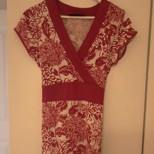 Rödvit blommig omlottklänning - Supersöt kortklänning med rödvitt blommönster och omlott-v-ringning. Klänningen har bred midja med knytband bak och korta ärmar. Perfekt för dig som gillar färg och mönster! Materialet känns mjukt och följsamt.