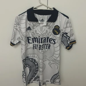 Real Madrid Vinicius Jr Adidas tröja - Säljer en Real Madrid fotbollströja från Adidas med Vinicius Jr och nummer 7 på ryggen. Tröjan är vit med grått drakmönster, svarta detaljer och guldinslag på ärmsluten. Klassisk pikékrage och klubbmärke på bröstet. Materialet är lätt och andas, perfekt för match eller träning.