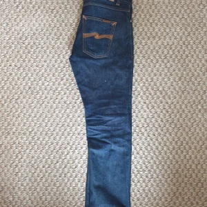 Mörkblå jeans från Nudie Jeans, W29 L30 - Snygga mörkblå jeans från Nudie Jeans med klassiska orange sömmar och ikonisk bakficka. Modellen har normal passform och raka ben. Tillverkade i slitstarkt bomullsjeans, perfekta för dig som gillar stilrena och tidlösa jeans.