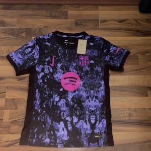 FC Barcelona Nike Dri-Fit fotbollströja - FC Barcelona Dri-Fit fotbollströja från Nike med svart och lila abstrakt mönster, rosa detaljer och klubbmärke på bröstet. Kortärmad modell i lätt och ventilerande material, perfekt för träning eller match. Spotify-logga i rosa framtill.”oanvänd”