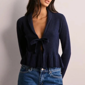 Mörkblå stickad kofta med knyt - Supersöt mörkblå stickad kofta med volangkant och knyt framtill. Djup v-ringning och långa ärmar ger en trendig look. Perfekt att styla med jeans för en chill vibe. Mjuk och skön, passar både till skolan och häng med vänner.