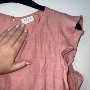 Rosa volangblus från VILA - Supersöt ljusrosa blus från VILA med volangdetaljer vid ärmhålen och nertill. Blusen är ärmlös och har en rund halsringning samt knapp i nacken. Perfekt för dig som gillar en feminin och luftig stil. Fåtal använd. Skulle passa S-L