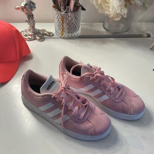Rosa Adidas sneakers i mocka - Snygga rosa Adidas sneakers med klassiska vita ränder på sidorna och vit sula. Skorna har snörning och är gjorda i mjuk mocka för en cool och sportig look. Perfekta för dig som vill sticka ut med färg och stil i vardagen.