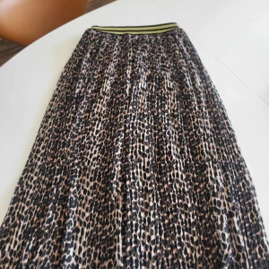 Leopardmönstrad långkjol från Staj - Snygg långkjol från Staj med leopardmönster i svart och beige. Kjolen har ett elastiskt midjeband med svarta och gula ränder som ger en sportig touch. Perfekt för dig som vill sticka ut med ett djurmönster och cool vibe.