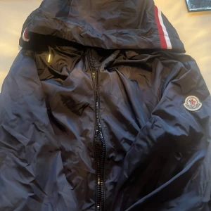 Svart vindjacka från Moncler - Snygg svart vindjacka från Moncler med huva och dragkedja framtill. Jackan har Moncler-logga på ärmen och coola röda och vita detaljer på huvan. Perfekt för dig som vill ha en stilren och sportig look.