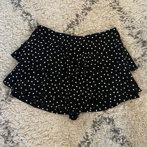 Prickiga volangshorts/kjol från River Island - Svarta shorts/kjol med vita prickar från River Island. De har flera lager volanger och en söt knytning framtill. Perfekta för dig som gillar en lekfull och trendig stil. Lätta och luftiga, passar grymt till sommaren.