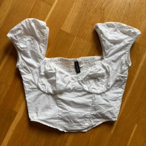 Vit korsettopp från HM - Supersöt vit korsettopp från HM med puffiga korta ärmar och smockad rygg. Vilket gör att den är lätt att sätta på sig och bekväm att bära. Toppen har markerade kupor och en croppad passform som ger en trendig siluett. Perfekt att ha på sig på en fest