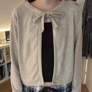 Beige kofta med knyt från Lager 157 - Supermjuk beige kofta från Lager 157 med långa ärmar och knytband framtill vid halsen. Koftan är i en ljus, melerad ton och har en enkel, clean look som passar perfekt till lager-på-lager. Materialet känns som bomull eller en mjuk syntetblandning. Använd ett fåtal gånger.