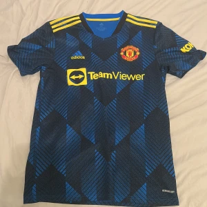 Manchester United bortatröja - Snygg Manchester United bortatröja från Adidas i svart och blått med gula detaljer. Tröjan har korta ärmar, geometriskt mönster och klubbmärke på bröstet. Tillverkad i lätt funktionsmaterial (AEROREADY) och har TeamViewer som sponsortryck. Pris kan diskuteras 