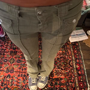 Olivgröna low waist utsvängda byxor från hm - Säljer ett par olivgröna cargopants med snygga sömdetaljer längs benen och fickor med lock och knapp framtill. Byxorna är low waisr och utsvänga, väldigt väldigt eftertraktade och populära!.