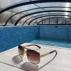 Guldiga Louis Vuitton solglasögon - Snygga solglasögon från Louis Vuitton med fyrkantiga guldiga metallbågar och bruna tonade glas. Skalmarna har gulddetaljer och mörkbruna inslag. Perfekt accessoar för att lyfta din stil vid poolen eller på stan.