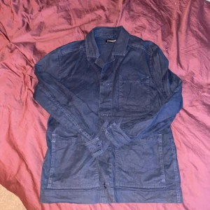 Blå overshirt från J.Lindeberg - Snygg blå overshirt från J.Lindeberg med klassisk krage och tre stora fickor framtill. Jackan är tillverkad i ett jeansliknande material och har knäppning framtill. Perfekt för lager-på-lager och en chill streetstyle.