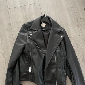 Svart skinnjacka med dragkedjor - Snygg svart skinnjacka från H&M med klassisk bikerstil. Jackan har silvriga dragkedjor, bred krage och coola detaljer. Perfekt för dig som vill ha en edgy look. Materialet är syntetiskt skinn och jackan har långa ärmar.