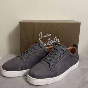 Grå mocka sneakers från Louboutin - Snygga grå sneakers i mocka från Christian Louboutin med klassisk vit sula och den ikoniska röda undersidan. Skorna har rund tå, snörning och diskreta detaljer. Insidan är fodrad i beige läder för extra komfort. Perfekta för dig som gillar stilrena och exklusiva sneakers.
