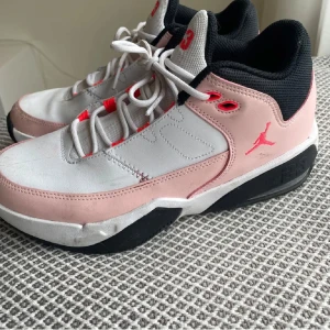Nike Air Jordan sneakers rosa/vit - Snygga Nike Air Jordan sneakers i vitt och ljusrosa med svarta detaljer och röda accenter. Skorna har klassisk snörning, rund tå och platt sula. Ovandelen är i syntet och skinn, med broderad Jumpman-logga och siffran 23 på plösen. Perfekta för dig som gillar streetstyle.