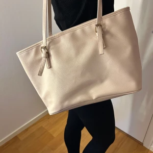 Beige axelväska med guldspännen - Stilren beige axelväska i syntetmaterial med två långa axelband och snygga guldiga spännen. Väskan är rymlig med flera innerfack, bland annat ett med dragkedja. Perfekt för dig som vill ha plats för allt du behöver under dagen.