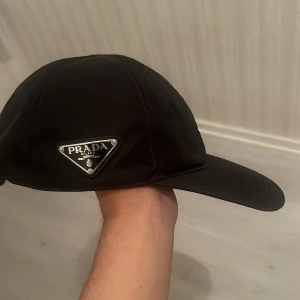 Svart keps från Prada i nylon - Svart keps från Prada i återvunnen nylon med klassisk böjd skärm och ikonisk metallogo på sidan. Snygg och stilren design som passar perfekt till streetwear eller en clean look. Justerbar baktill för bästa passform. 