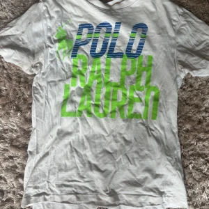 Vit t-shirt Polo Ralph Lauren - Vit t-shirt från Polo Ralph Lauren med stor blå och grön logga framtill. Klassisk rund hals och korta ärmar. Tillverkad i mjuk bomull, perfekt för en chill och stilren look. Snyggt tryck som sticker ut och ger en sportig vibe. Köpt på sellpy och är väldigt använd🤍
