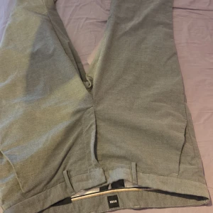 Grå kostymbyxor från BOSS - Snygga grå kostymbyxor från BOSS med klassisk rak passform och bälteshällor. Byxorna har diskreta bakfickor och är tillverkade i ett mjukt tyg som känns bekvämt. Perfekta för en clean och stilren look. Liten fläk på baksidan som säkert går bort vid nästa tvätt och som knappt syns. 