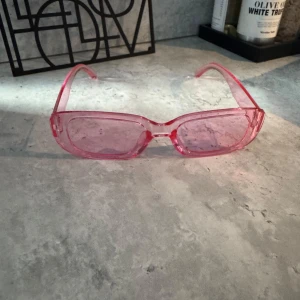 Rosa rektangulära solglasögon - Snygga solglasögon med rektangulära bågar i transparent rosa plast. Glasen är också rosa och ger en cool vibe. Perfekta för dig som vill sticka ut och addera färg till din stil. Passar till alla som gillar trendiga accessoarer.