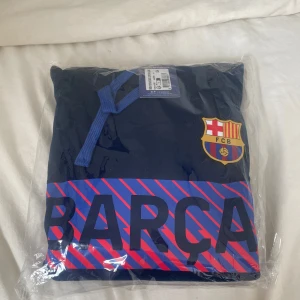 FC Barcelona hoodie marinblå S - Marinblå hoodie från FC Barcelona med klubbens emblem på bröstet och stor BARÇA-text i blått och rött framtill. Tröjan har huva med blå snören och är perfekt för dig som älskar fotboll och vill visa ditt stöd för Barça. Helt oöppnad och aldrig använd.