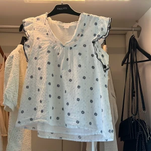 Vit blommig blus från Lindex - Supersöt vit blus från Lindex med små mörkblå blommor och volangdetaljer vid ärmhålen. Blusen är ärmlös med v-ringning och har svarta vågiga kanter på volangerna. Perfekt för en fräsch och somrig look!