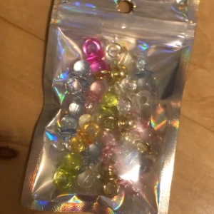 Färgglada napp-berlocker till smyckestillverkning - En påse med 23 st mixade napp-berlocker i olika färger som rosa, blå, gul, grön och guld. Berlockerna är runda, genomskinliga och ser ut som nappar, perfekta för att skapa egna armband, halsband eller örhängen. Kommer i en holografisk förpackning som ger extra cool känsla. 