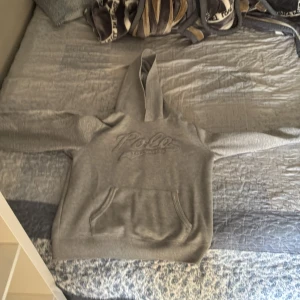 Grå hoodie från Polo Ralph Lauren - Snygg grå hoodie från Polo Ralph Lauren med broderad logga på bröstet. Klassisk känguruficka framtill och huva. Perfekt för dig som gillar en enkel och stilren look. Mjuk och skön bomullsblandning, passar till många outfits.