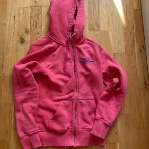 Rosa hoodie Peak Performance S - Rosa hoodie från Peak Performance med blå broderad logga på bröstet. Tröjan har dragkedja, huva med snörning och två fickor framtill. Den har en liten fläck vid ena armen men den syns inte tydligt. 
