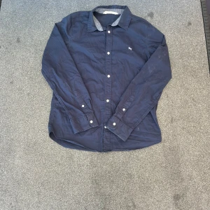 Mörkblå bomullsskjorta med knappar - Snygg mörkblå skjorta i bomull med klassisk krage och vita knappar. Liten broderad logga på bröstet och kontrasttyg på insidan av kragen. Perfekt för en stilren och avslappnad look.