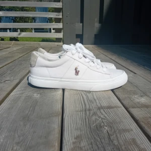 Vita sneakers från Polo Ralph Lauren - Fräscha vita sneakers från Polo Ralph Lauren med klassisk logga på sidan och POLO-tryck på hälen. Skorna har snörning, ovandel i bomullstyg och detaljer i mocka på hälen. Perfekta för en clean och stilren look. Det finns en liten missfärgning se sista bilden. Finns fler bilder vid behov.