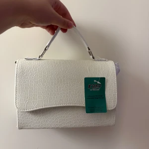 Vit handväska/axelväska  från Kobo med struktur - Snygg vit handväska/axelväska med vitt band från Kobo med croco-mönstrad struktur och stilrent handtag. Väskan har en klassisk rektangulär form och är tillverkad i syntetmaterial. Perfekt för dig som vill ha en clean och trendig accessoar till din outfit.