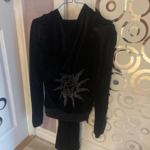 Svart sammetshoodie med matchande byxor  - Svart hoodie i mjuk sammet från Isle of You. Tröjan har dragkedja framtill, huva och coola strassdetaljer med sol- och ansiktsmotiv på ryggen och benet. Långa ärmar och snörning i midjan ger en snygg och bekväm passform, med matchande byxor. storlek xs-s 